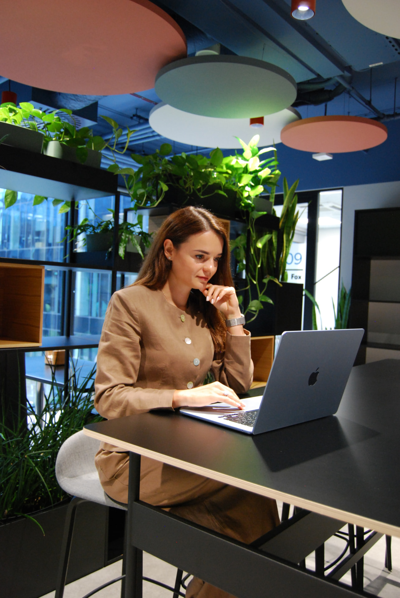 Angelica working on her laptop in a modern office with plants and colourful ceiling lights